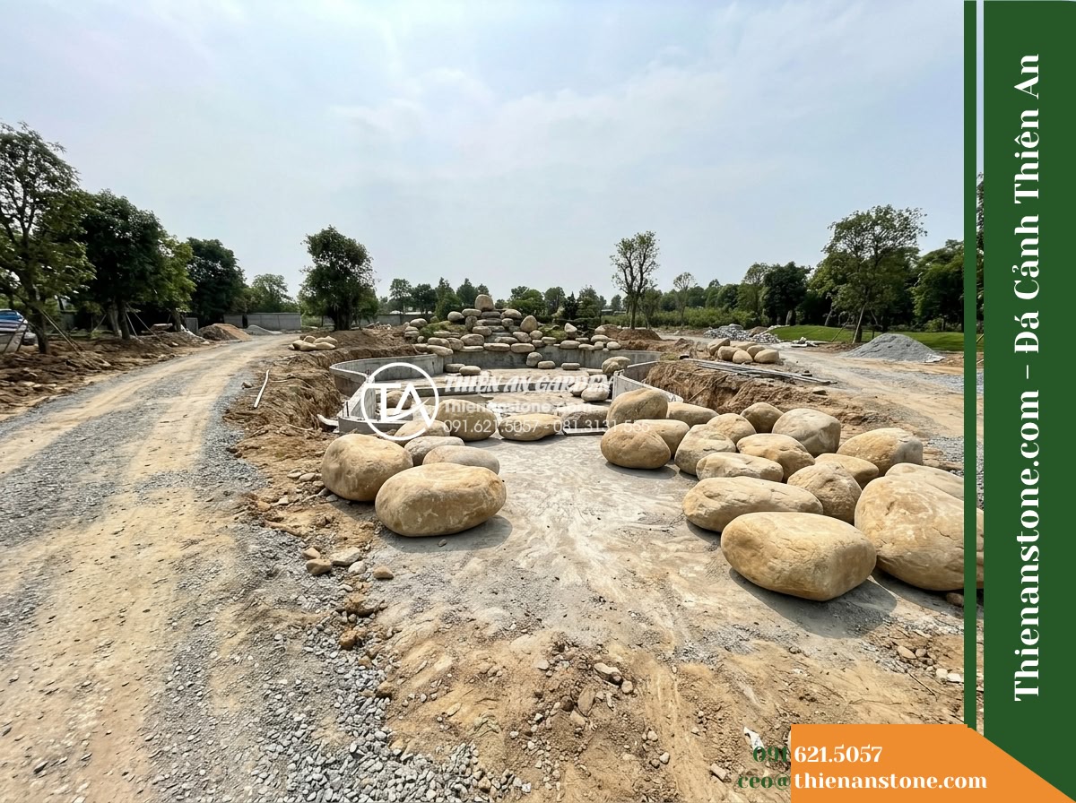 Đá Niên Thủy (Cuội đại 80–150cm / ~0.5–2 tấn) | Chuyên làm gò đất thấp, bãi đá ngầm chặn dòng dưới chân thác non bộ. - Hình 9 Đá Niên Thủy (Cuội đại 80–150cm / ~0.5–2 tấn) | Chuyên làm gò đất thấp, bãi đá ngầm chặn dòng dưới chân thác non bộ. - Wide site view of an outdoor landscape area showing access paths, staging ground
