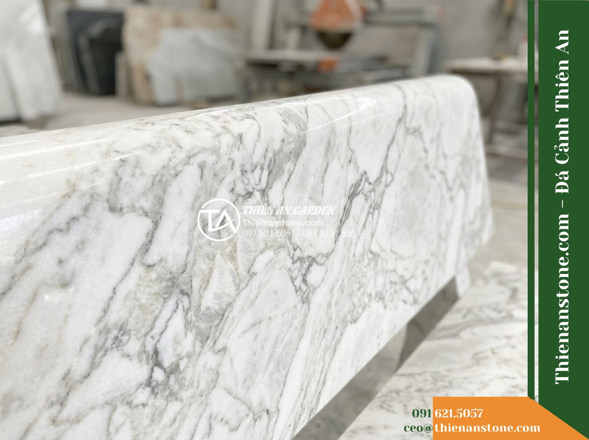 Ghế Đá Công Viên Không Tựa Liền Khối Thiên Kim 160x50x45cm - Đá Vàng Cẩm Thạch - Hình 2 Ghế Đá Công Viên Không Tựa Liền Khối Thiên Kim 160x50x45cm - Đá Vàng Cẩm Thạch - A beautifully crafted golden marble stone bench under a large banyan tree, dappl